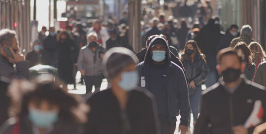 People wearing masks walk along a crowded city street.