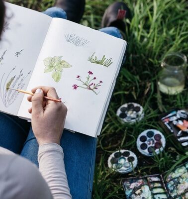 A person sitting on the ground with watercolours and painting in a journaling book