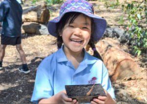 Small child holding up a seedling smiling at the camera. They are wearing a bucket hat and blue tshirt.